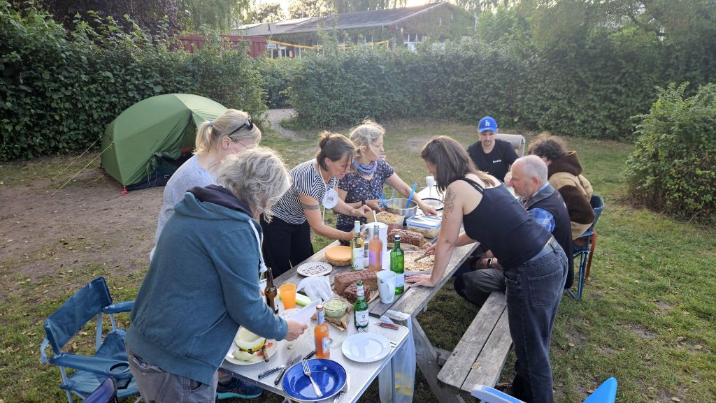 Acht Menschen, die auf einem Camping-Platz gemeinsam Essen zubereiten. Die Menschen stehen bzw. sitzen an eniem Tisch, im Hintergrund steht ein Zelt.